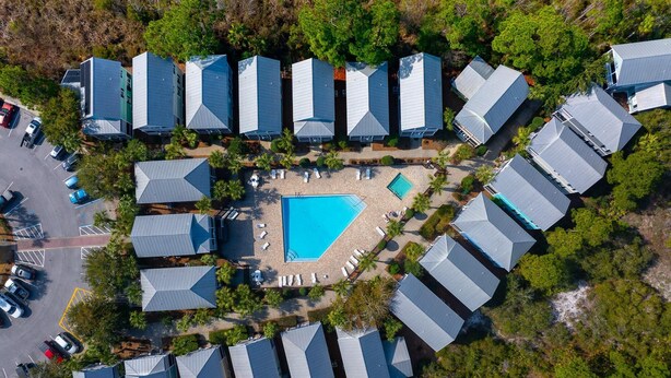 Eine Vogelperspektive zeigt einen Urlaubsort mit mehreren bungalowsartigen Gebäuden, die um einen zentralen Pool angeordnet sind.