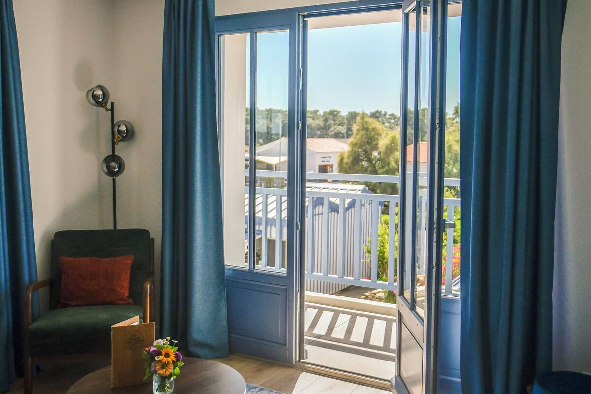 A modern living room with a yellow sofa, a green armchair, and a small round table with a vase of flowers. A balcony with blue curtains is visible through the open door.