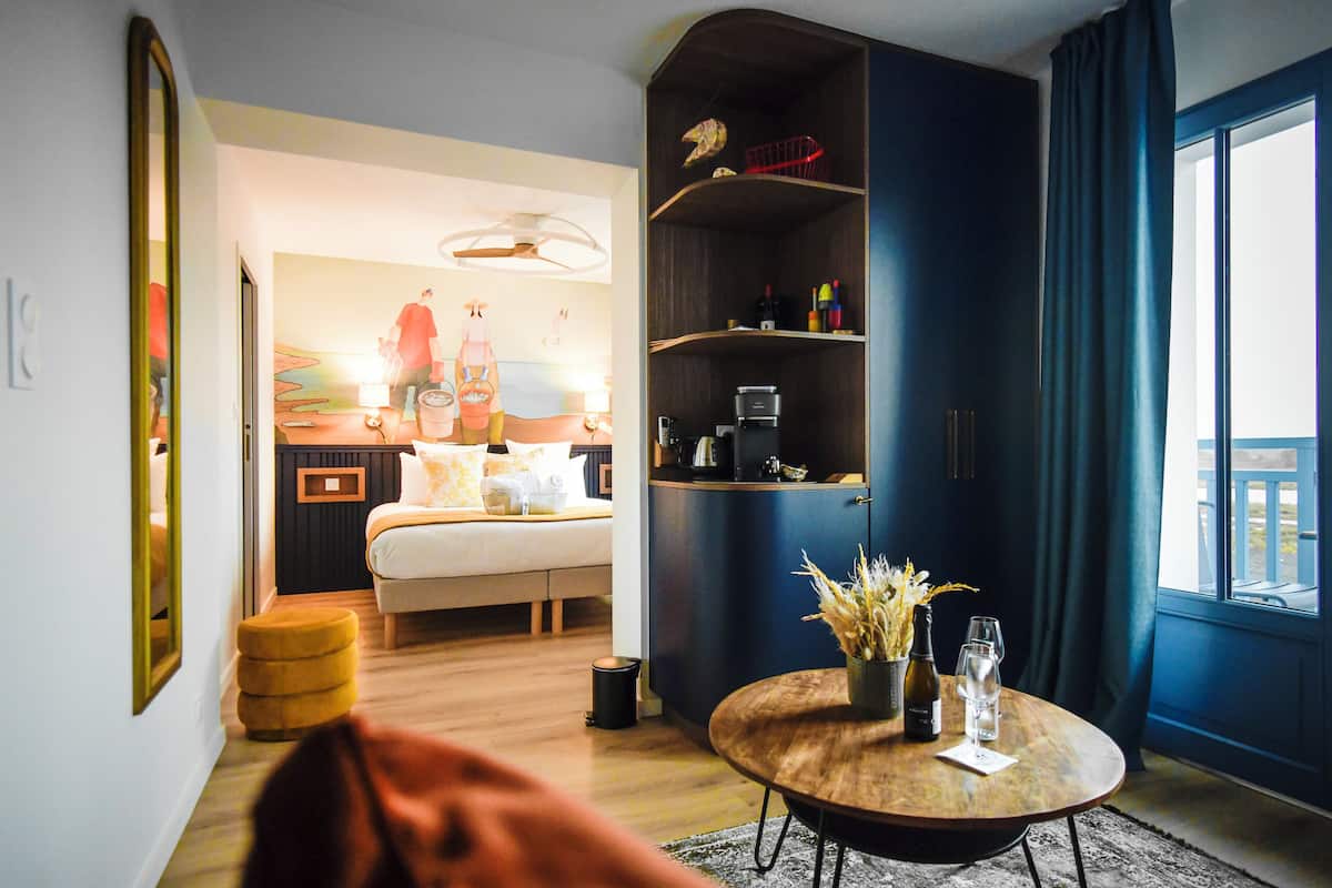 A modern hotel room with a bed, a round wooden coffee table with a vase and glasses, a bookshelf, and a large window with curtains.