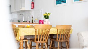 A dining area with a yellow tablecloth, wooden chairs, a vase with flowers, and a kitchen in the background.