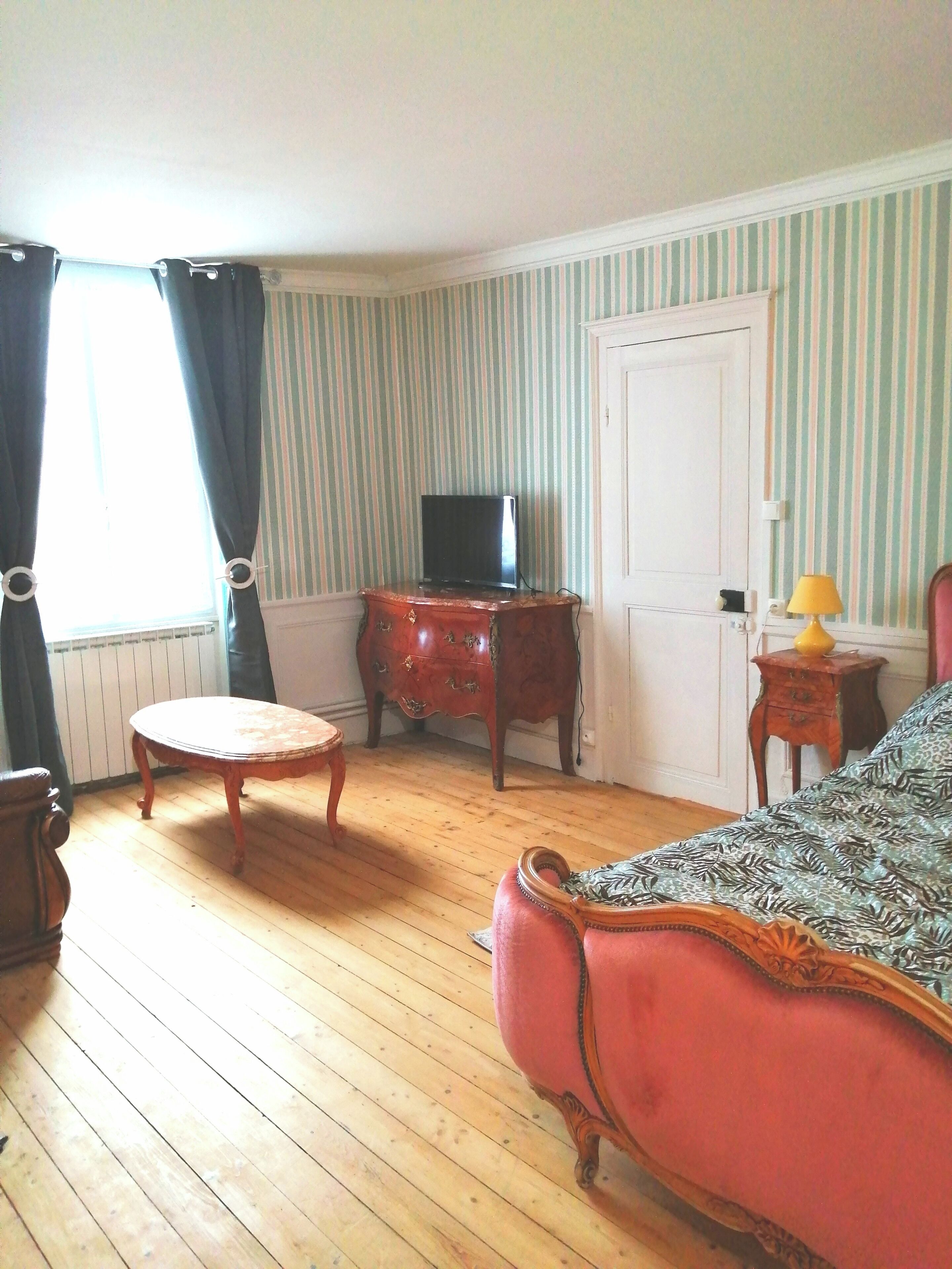 A room with a wooden floor, a red upholstered sofa, a small round table, a television on a wooden cabinet, a door, and a window with curtains.