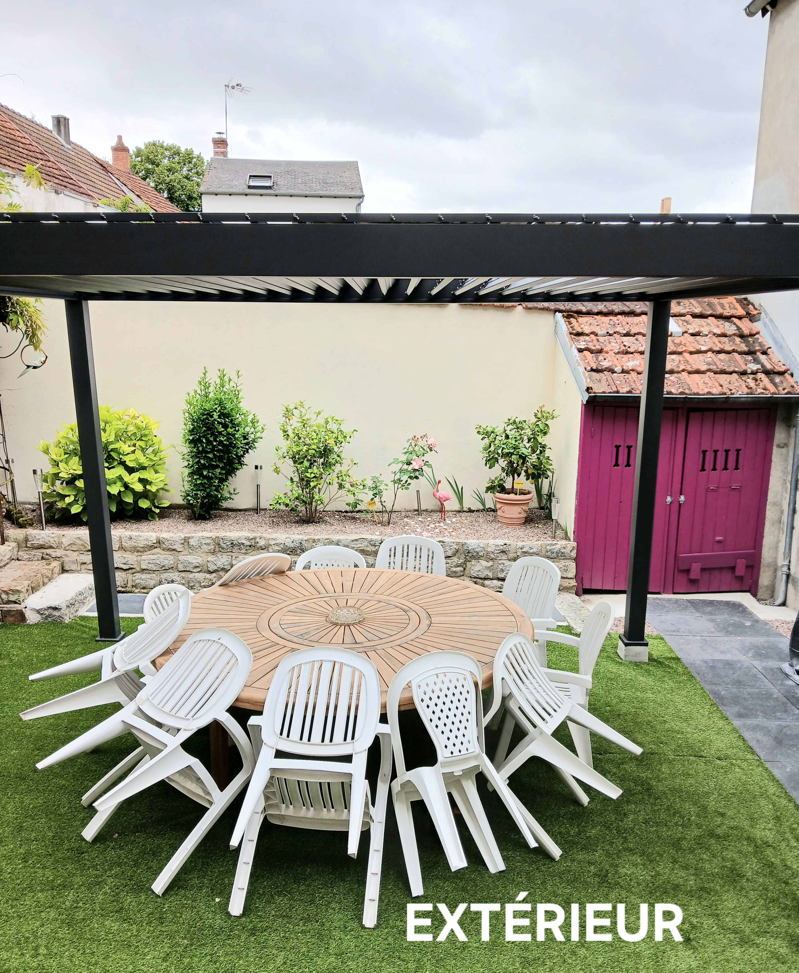 Een buitenpatio met een ronde houten tafel en witte plastic stoelen, omgeven door kunstgras.