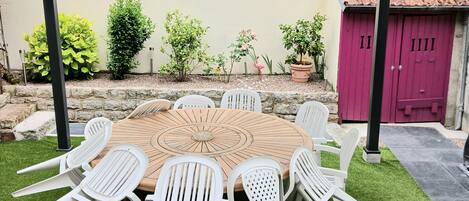 Een buitenpatio met een ronde houten tafel en witte plastic stoelen, omgeven door kunstgras.