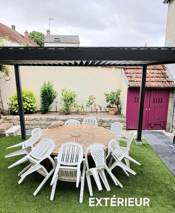 Eine Terrasse im Freien mit einem runden Holztisch und weißen Plastikstühlen, umgeben von grünem Kunstgrün.