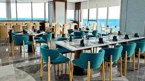 A restaurant with teal chairs and wooden legs, tables set for dining, and large windows offering a view of the ocean.