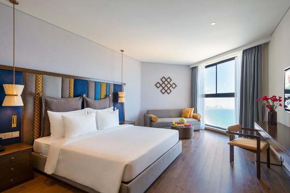 A hotel room with a large bed, a sofa, a small table, and a view of the sea.