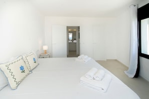 A hotel room with a bed, pillows, a nightstand, a lamp, a bathroom visible through an open door, and a window with curtains.