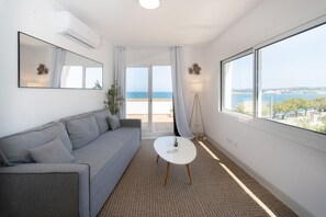 A modern room with a grey sofa, a small white table, and a large window offering a view of the sea.