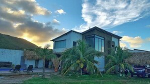 Una casa de dos pisos con un ventanal grande, techo de paja y zona de piscina.