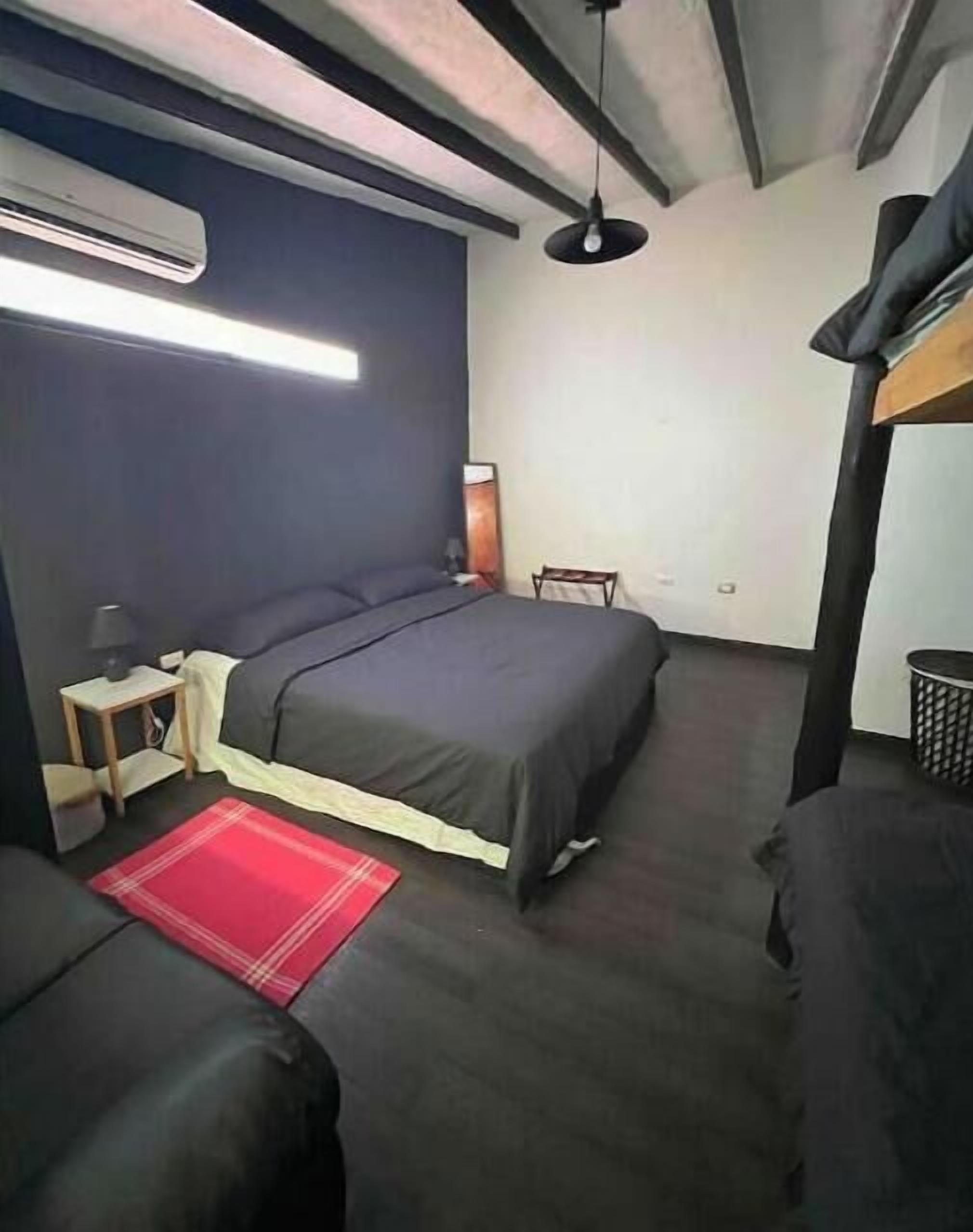 Une chambre à coucher de taille réduite, avec un lit simple, une petite table de chevet, un tapis rouge, un luminaire suspendu et un lit superposé dans un coin.