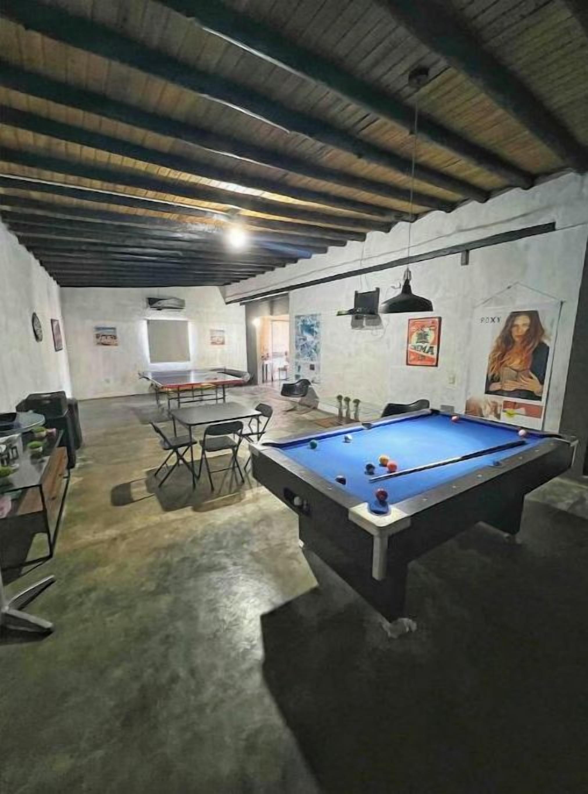 A pool table in a room with a concrete floor and a high ceiling. There is a table and chairs in the background.