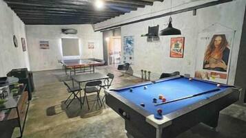 A pool table in a room with a concrete floor and a high ceiling. There is a table and chairs in the background.
