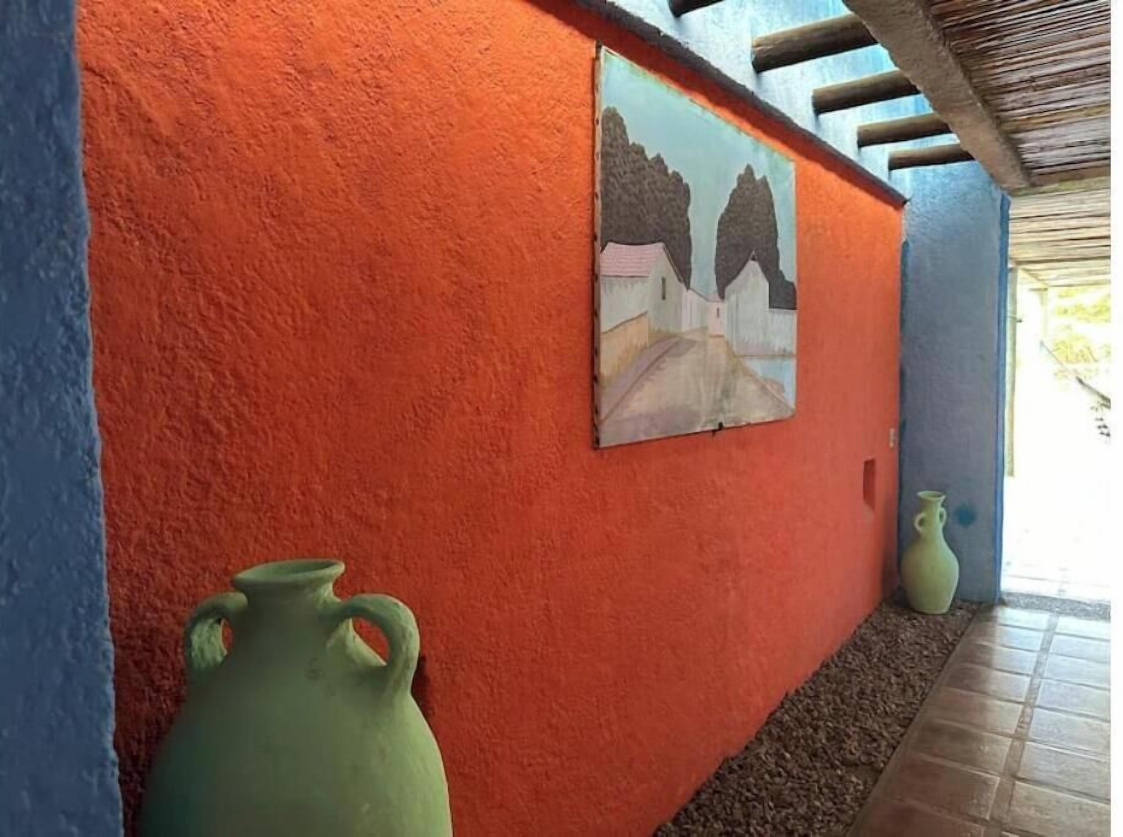 A corridor with a textured orange wall, a painting of houses, and two green vases.