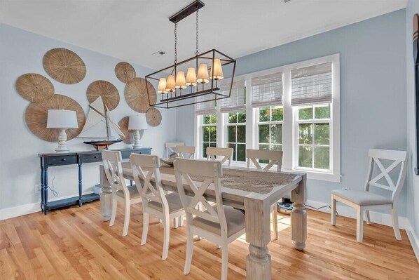 A dining area with a wooden table and chairs, a chandelier, and decorative wall art. - 3BR + Loft, Walk to Beach, Porch Swing & DOGS OK (Emerald Isle)