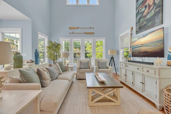 A spacious living room with a large sofa, two armchairs, a coffee table, and a cabinet with a television. There are windows with white frames and a view of greenery outside.
