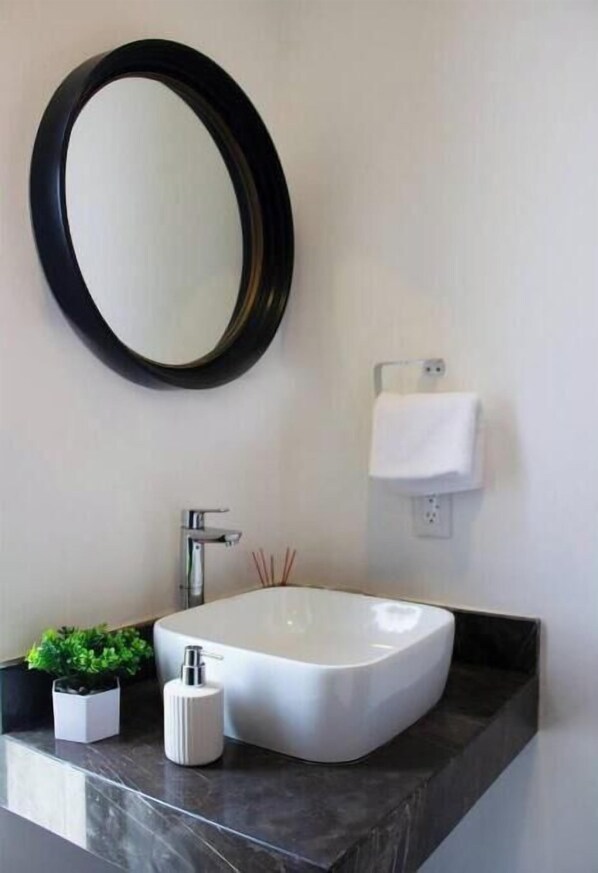 A modern bathroom with a large oval mirror, a white sink, a black countertop, a small potted plant, and a white towel dispenser.