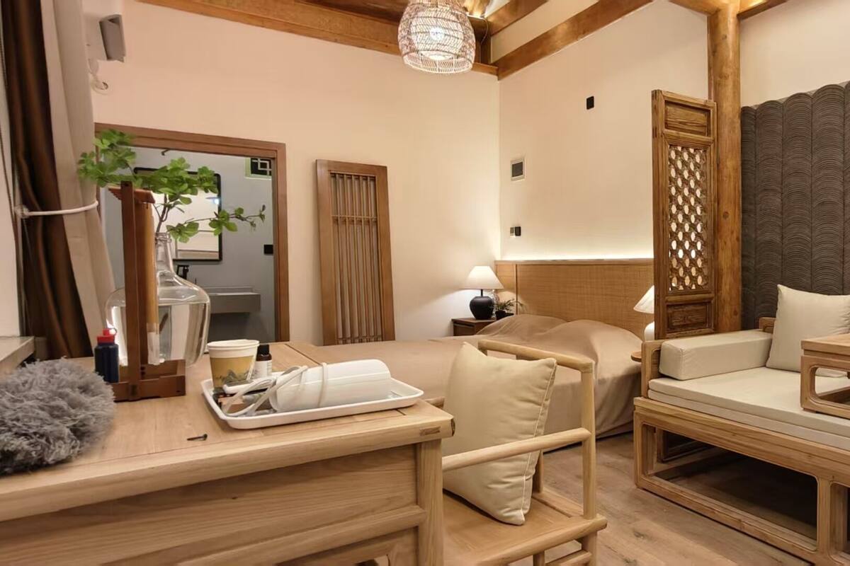A cozy room with a wooden bed, a small wooden table, a chair, and a large cushioned bench.