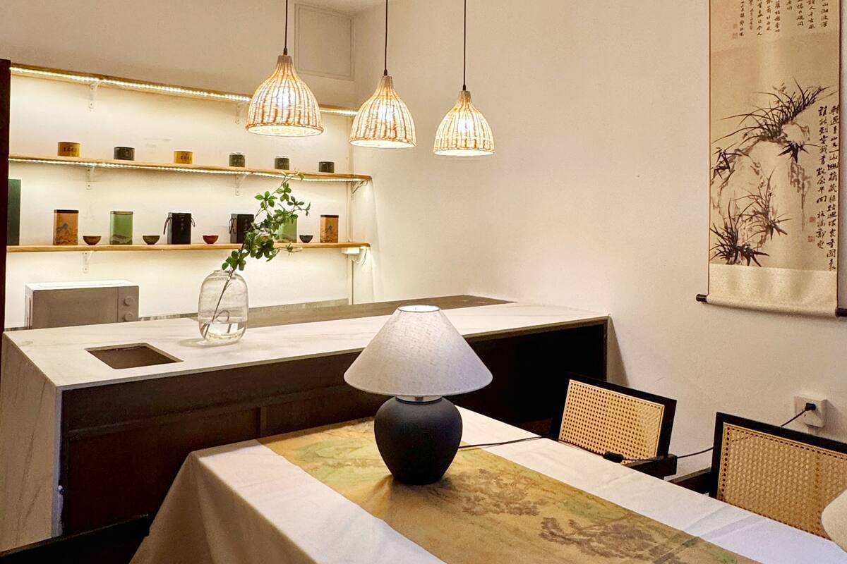A dining area with a table, chairs, a lamp, hanging lights, a vase with a plant, and a wall with a hanging scroll.