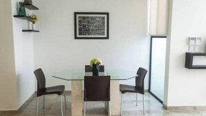 A modern dining area with a glass table surrounded by four chairs, a framed artwork on the wall, and a small shelf with decorative items.