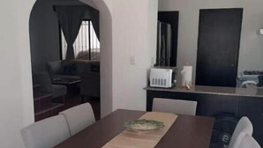 Uma área de jantar com uma mesa retangular, cadeiras brancas e um jogo americano marrom. Há uma cozinha ao fundo, com um micro-ondas e um armário preto.