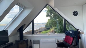 Et værelse med et stort vindue der ud til en bro og vandet, et fjernsyn, en pejs og en lænestol med en sportstrøje.