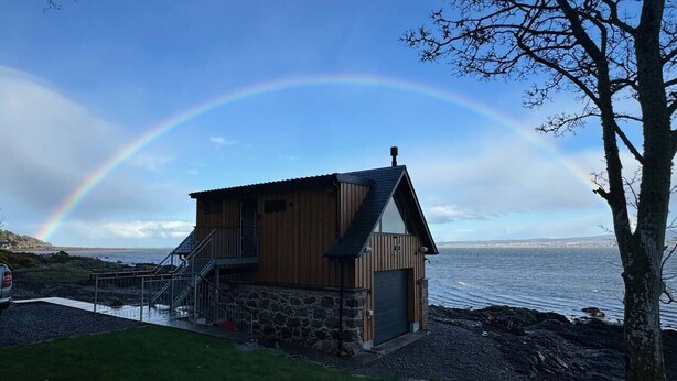 En træhytte med stengrunding ved en stenet kystlinje, en trappe op til en terrasse, og udsigt til havet.