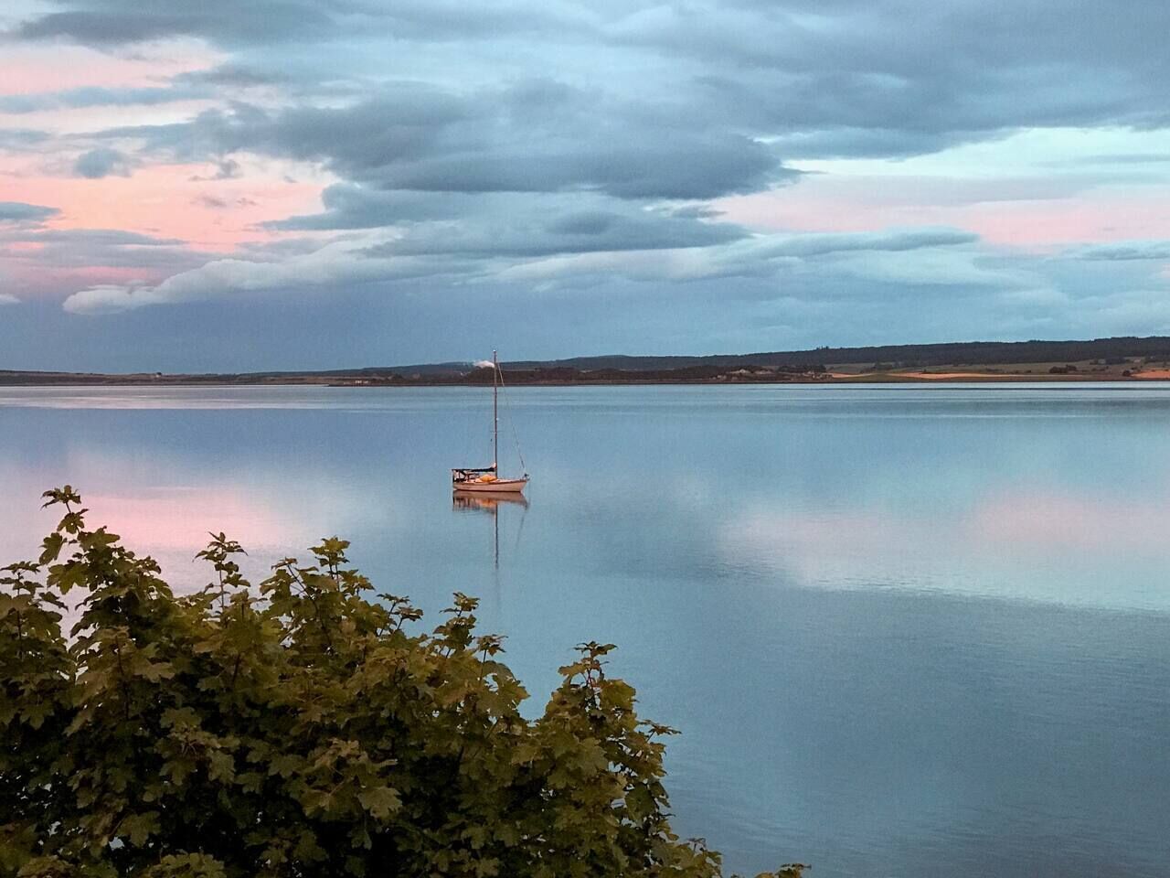 Een rustig meer met een zeilboot in het midden, omringd door bomen en een bewolkte lucht.