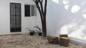 A white building with a window and a door, surrounded by gravel, potted plants, and wooden stumps.