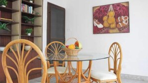 A dining area with a glass table surrounded by wicker chairs, a bookshelf with books and plants, and a painting on the wall.