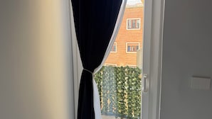 Une fenêtre avec des rideaux noirs, une vue sur un bâtiment en briques et une clôture verte à l’extérieur.