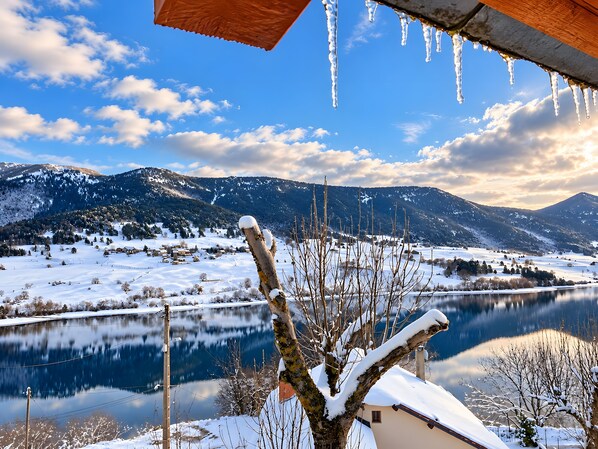 A snowy landscape with a lake, mountains, and a house with a sloped roof.