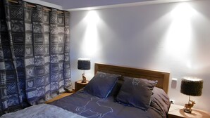 Een slaapkamer met een bed, twee nachtkastjes, een lamp, een plaid en een gordijn met een patroon.