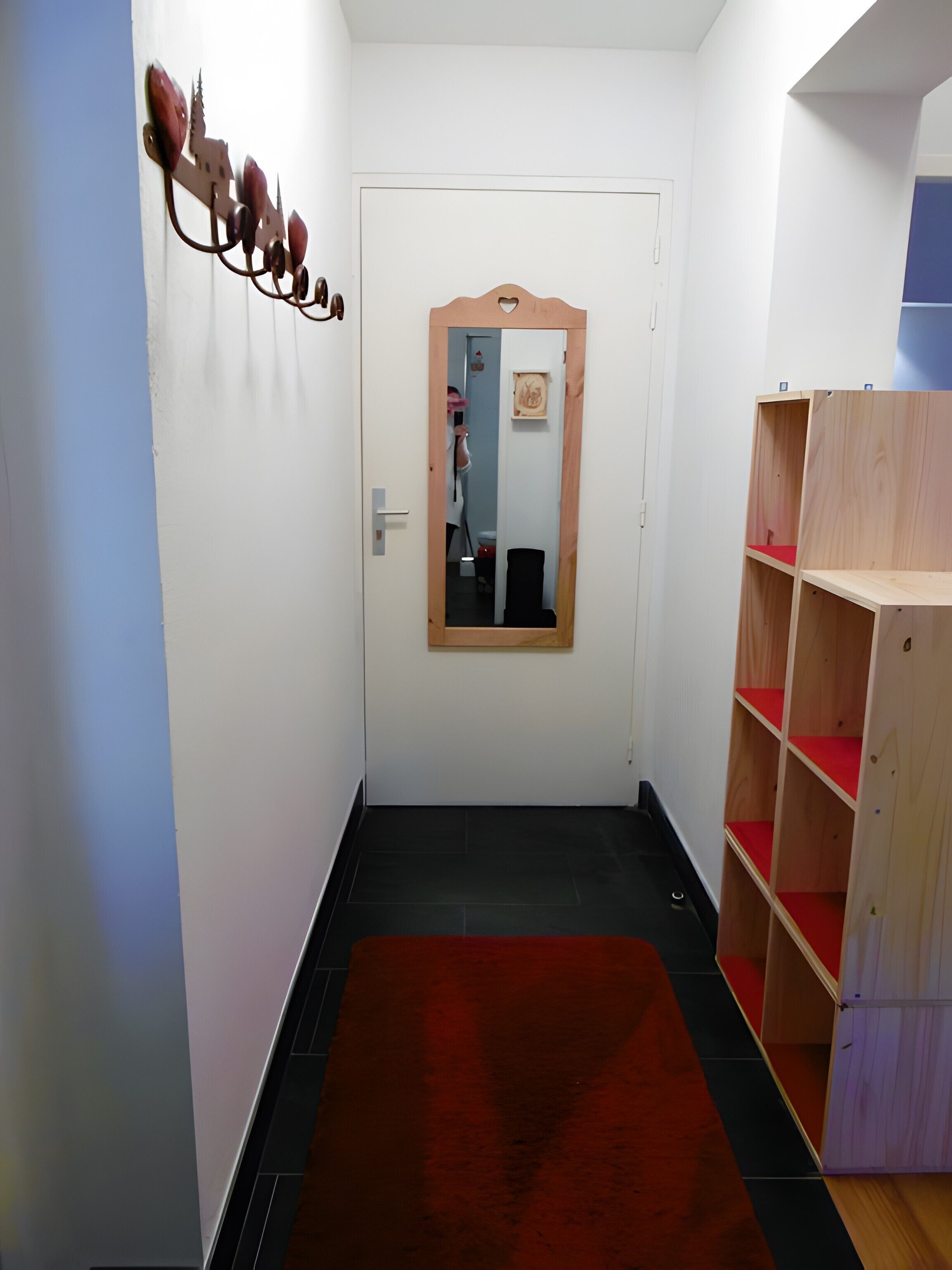 Un corridoio con tappeto rosso, una porta in legno e uno specchio che riflette una stanza con una mensola rossa.