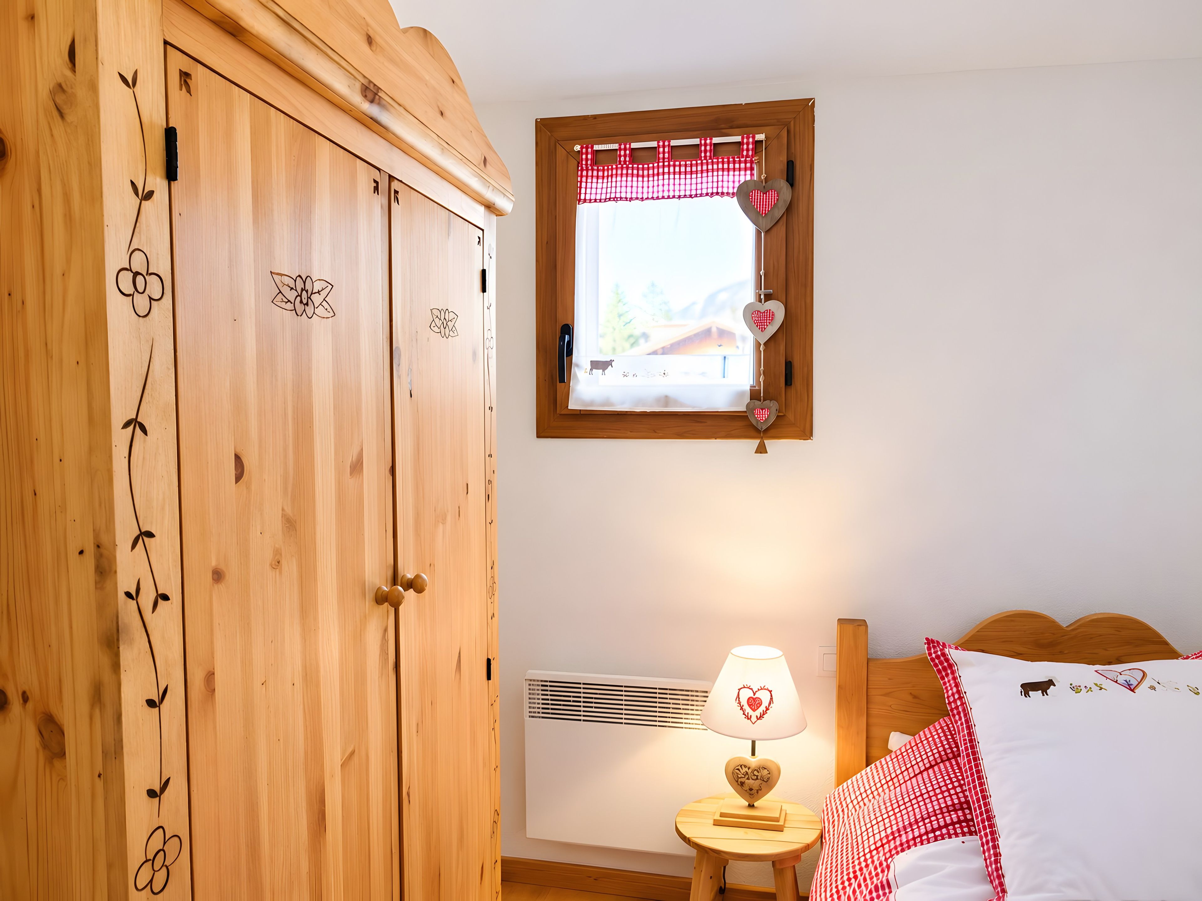 A bedroom with a wooden wardrobe, a bedside table with a lamp, a window with a checkered curtain, and a bed with patterned bedding.