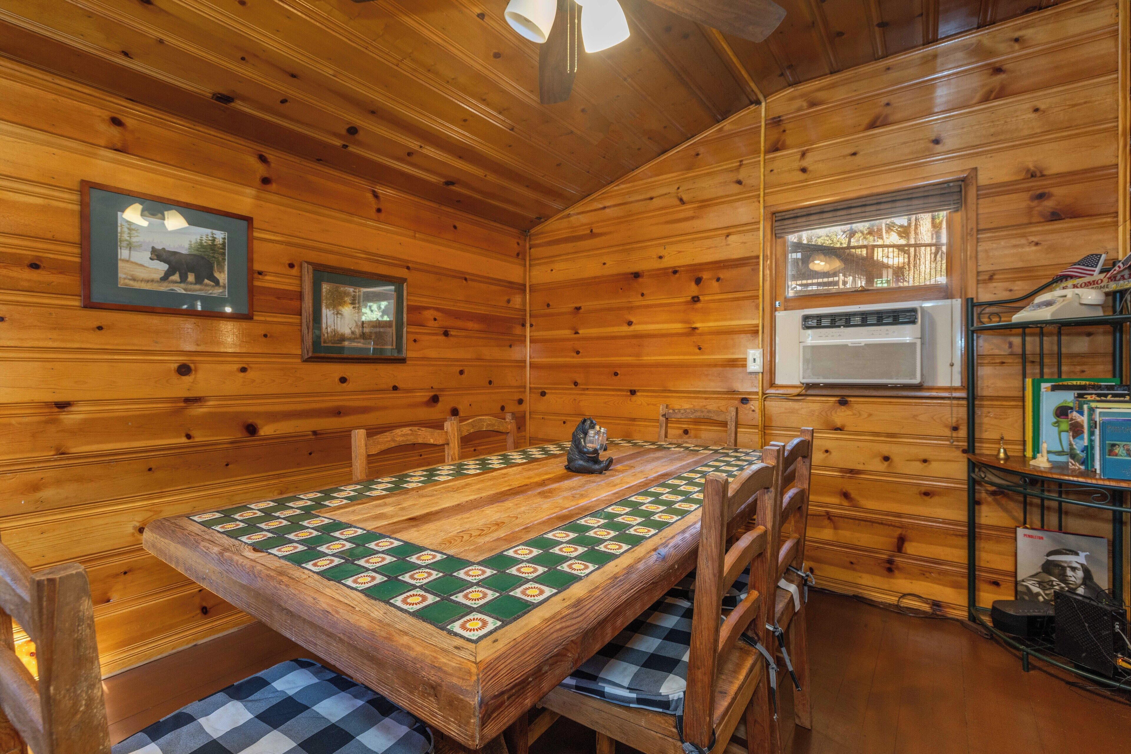 L'intérieur d'un chalet en bois, avec une table à manger, des chaises, des tableaux encadrés, une fenêtre et une étagère contenant des livres et une petite statue.