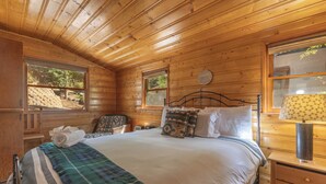 A wooden cabin bedroom with a bed, pillows, a nightstand, and a window. - True Big Bear Lake Cabin with jacuzzi (Big Bear Lake)