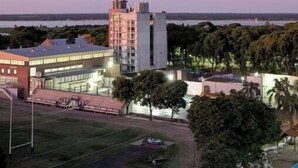 Un edificio de varios pisos con techo plano y grandes ventanales, rodeado de árboles y un campo deportivo.