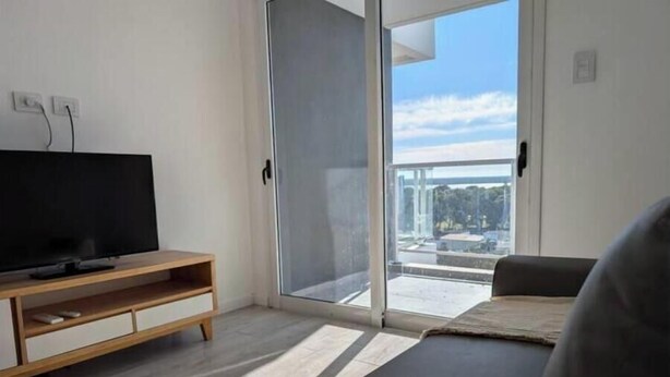 Una sala moderna con una televisión de pantalla plana sobre un soporte de madera, una puerta de vidrio corrediza que da a un balcón y un sofá gris.