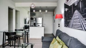 Un salon moderne avec un canapé gris, une table basse en verre et un coin repas avec des chaises noires et une table en verre. On aperçoit au fond une cuisine avec des meubles blancs et une lampe rouge.