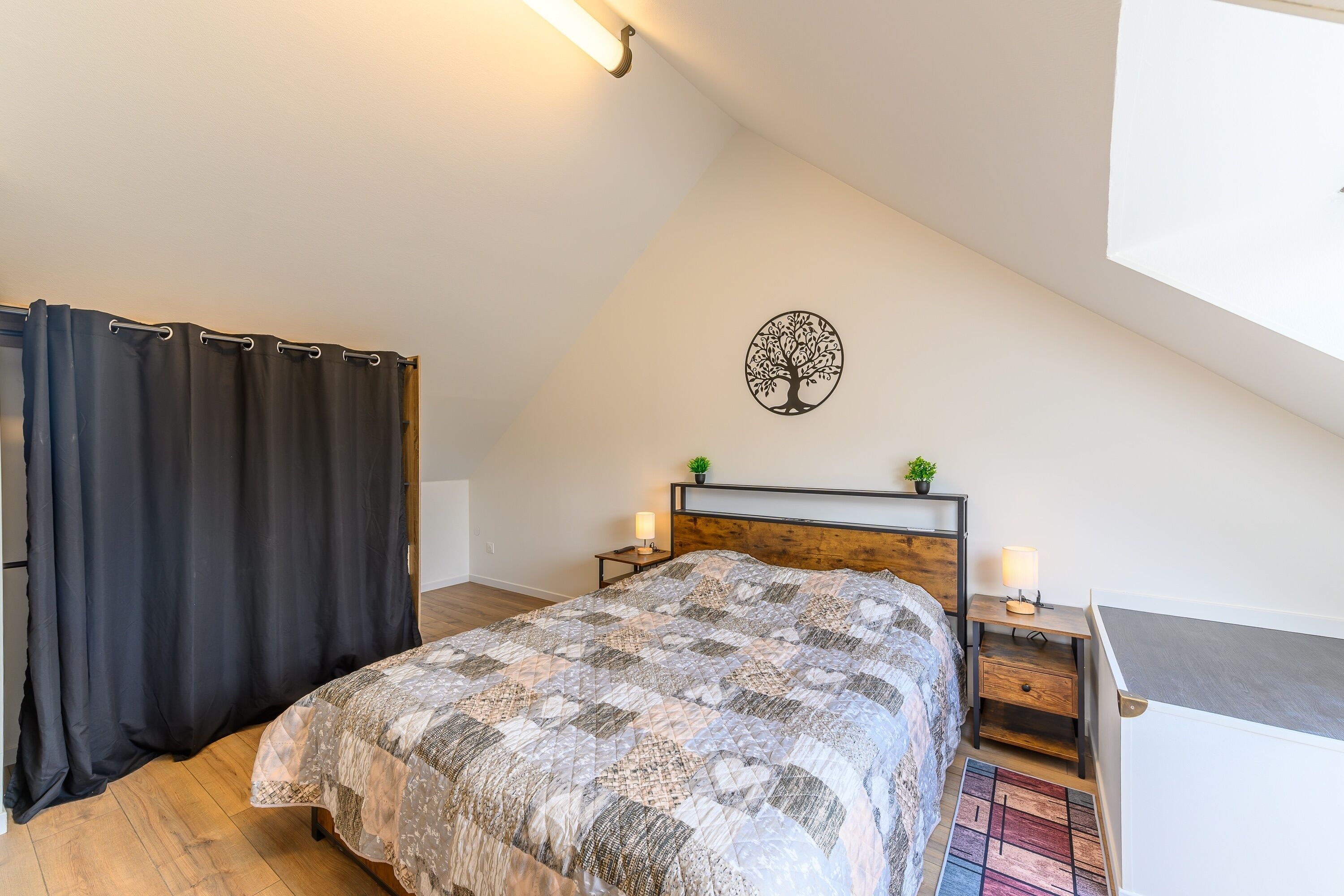 Un dormitorio con techo inclinado, una cama con colcha estampada, una mesita de noche con lámpara, una planta pequeña y una obra de arte en la pared con un árbol.