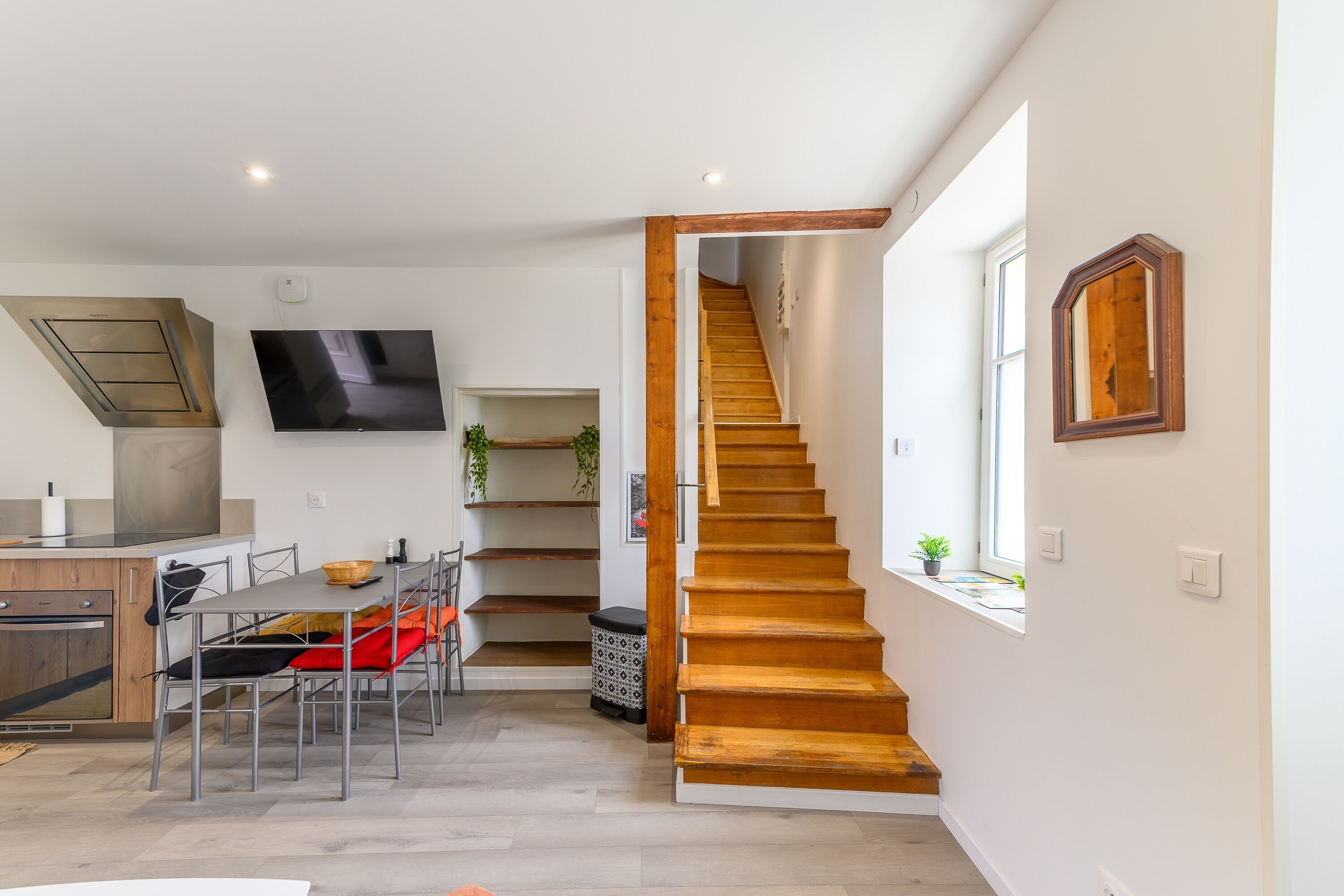 Une cuisine moderne dotée d’un four encastré, un coin repas avec une table et des chaises, un escalier en bois et un téléviseur fixé au mur.