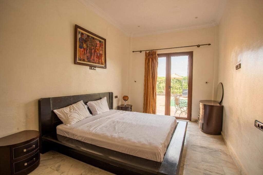 Une chambre avec un grand lit, une table de chevet, une commode, un tableau au mur et une porte-fenêtre coulissante donnant sur un espace extérieur.