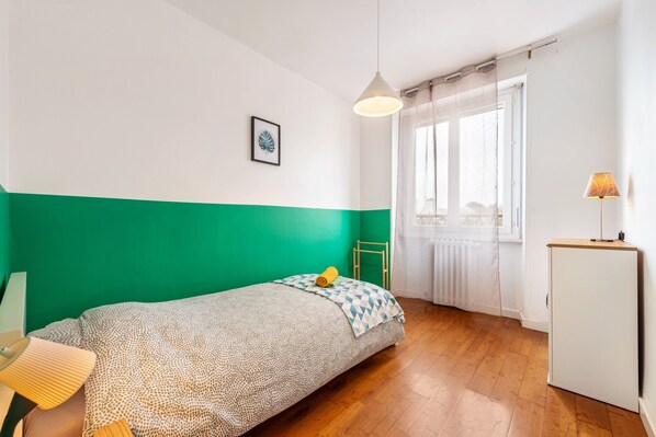 Un dormitorio con cama, escritorio, lámpara, radiador y ventana con cortinas.