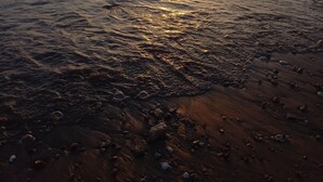 Una spiaggia con il sole che tramonta all'orizzonte, le onde che lambiscono dolcemente la riva e ciottoli sparsi.