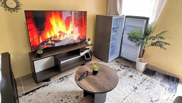 A living room with a flat-screen TV displaying a fireplace, a small round table with a remote, and a potted plant. - Rossyln haven studio (Nairobi)