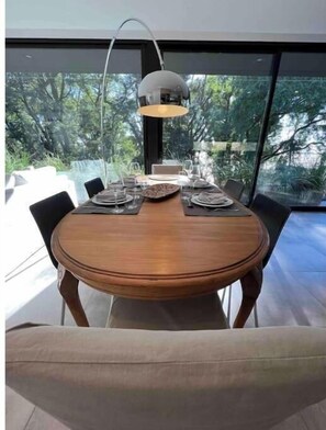 A dining table set for four with a modern pendant light, surrounded by chairs, in a room with large windows offering a view of greenery outside.