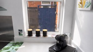 A kitchen with a coffee maker on the counter, a window with a view of a blue gate, and a small shelf with cups. - Entire Excellent Property on Mountcastle Road, Leicester (Leicester)