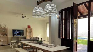 Un salon spacieux avec une table à manger en bois, des bancs et un téléviseur. On y trouve deux luminaires suspendus et un ventilateur de plafond.