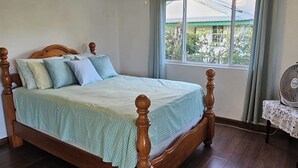 Un dormitorio con una cama de madera, un ventilador de techo, una ventana con cortinas y un ventilador de mesa.