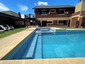 Piscina retangular com borda azulejada, rodeada por um deck de concreto. Há espreguiçadeiras e um espaço coberto com varanda e escada que leva a um segundo andar.